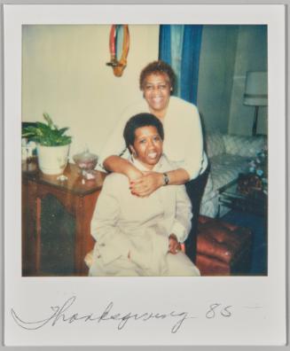 Thanksgiving '85 [Woman sitting on chair and smiling while other woman hugs her around the shoulders from behind]
