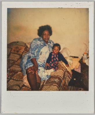 [Woman in blue dress and round glasses laying on bed next to baby]
