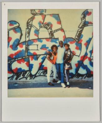 [Young man posing with two young women in front of a graffiti wall]
