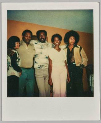 [Group posing in front of yellow wall, man on right has an afro]
