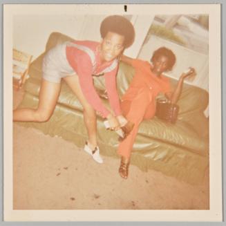 [Woman in overalls pulling high heel off of friends foot sitting on couch]
