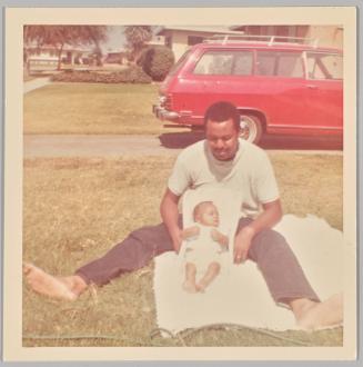[Man sitting on white towel on lawn with baby between his legs sleeping in car seat]
