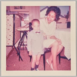 [Little boy wearing a short-suit and bowtie, standing next to woman sitting at table]
