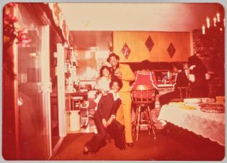 [Two men and woman posing at a house party with people seated at bar on the right side]
