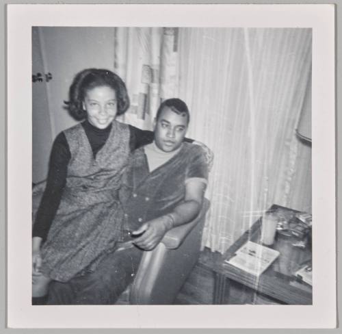 [Woman sitting on man's lap in front of curtained window]
