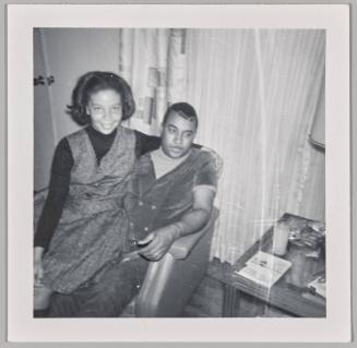 [Woman sitting on man's lap in front of curtained window]
