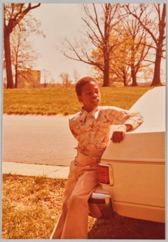 [Little boy dressed formally sitting on bumper of car beside road]
