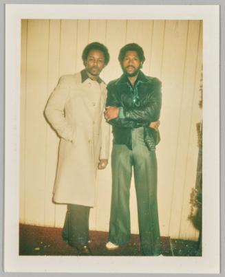 [Two men posing in front of white wall wearing suit pants and jackets]
