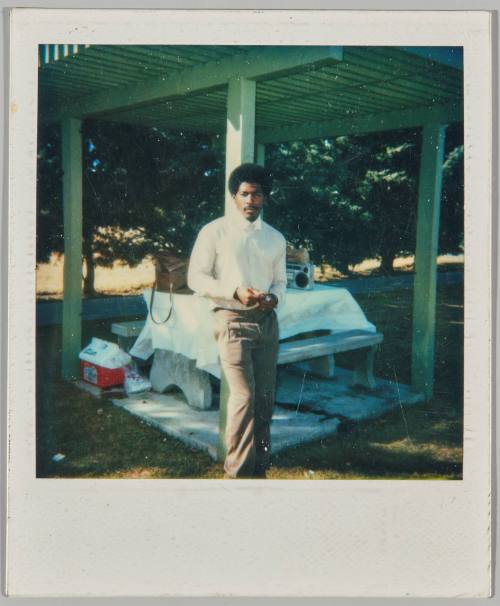 [Man leaning against wooden beam in front of picnic table]
