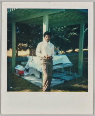 [Man leaning against wooden beam in front of picnic table]

