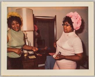 [Two women in pastel decorative hats in front of mirror and lamp]
