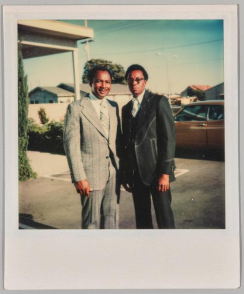 [Two men in suits posing in parking lot]
