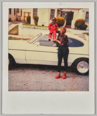 [Woman holding up little boy in red on white car]

