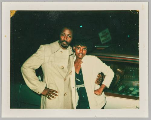 [Man and woman posing with arms around each other and hands on hip in front of car]
