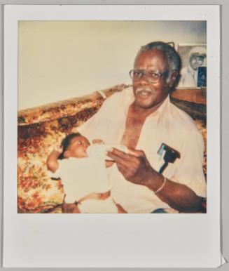 [Man feeding baby in arms with bottle on couch]
