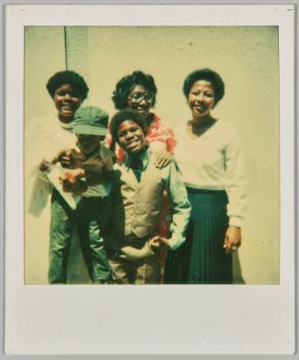 [Three women with two boys posing in formal outfits against concrete wall]
