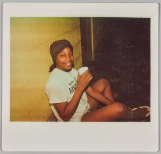[Young woman wearing durag sitting against cement wall, holding cup]
