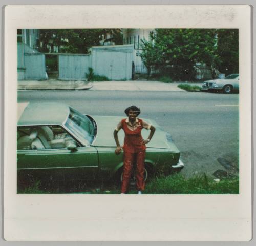 [Woman in red suit leaning on hood of green car]
