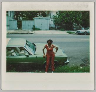 [Woman in red suit leaning on hood of green car]
