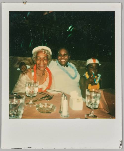 [Elderly woman, woman and child sitting at table with party necklaces and blowers]
