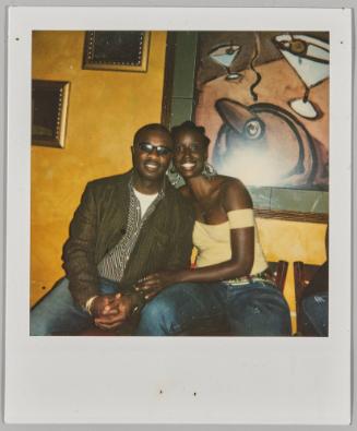 [Man and woman holding hands seated with painting in background]
