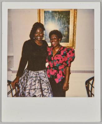 [Two women dressed formally posing in front of embellished framed painting]
