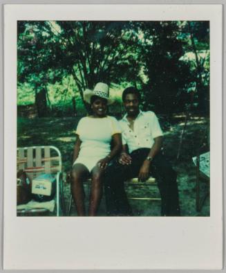 [Woman in straw hat sitting with man on folding chairs outdoors]
