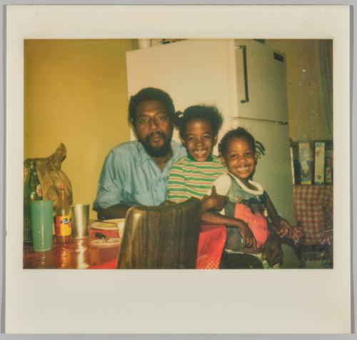 [Two smiling girls sitting on mans lap at dining table in front of fridge]
