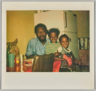 [Two smiling girls sitting on mans lap at dining table in front of fridge]
