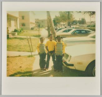 [Boy and two girls with matching outfits on sidewalk next to parked cars]

