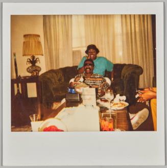 [Woman making peace sign, man seated at her feet eating meal at coffee table]
