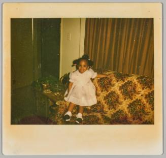 [Little girl in pink dress with braids sitting on arm of patterned couch]
