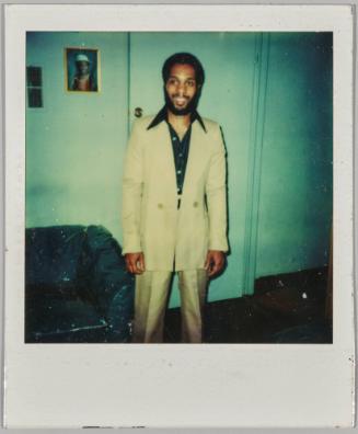 [Man in pale yellow suit smiling in front of door]
