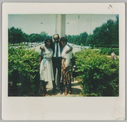[Man and two women dressed formally standing in front of parking lot]
