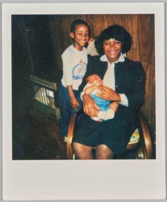 [Woman in rocking chair holding baby with boy at her side]
