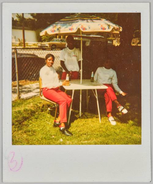 [Three young women in matching outfits seated around outdoor table drinking beer]
