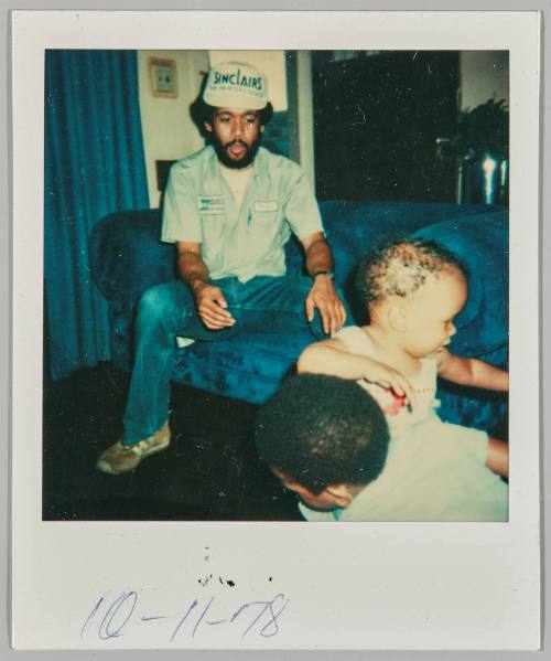10-11-78 [Man in blue work uniform sitting on a blue couch extends hand to toddler]