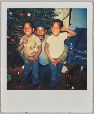 [Man with moustache and hat crouching in between two young boys smiling, Christmas Tree behind them]