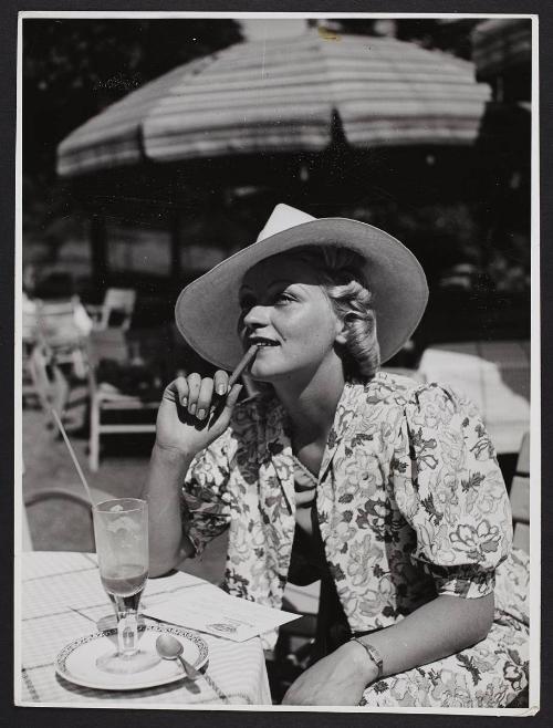 Woman in a floral dress and hat, chewing a pencil.