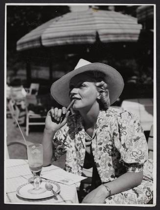Woman in a floral dress and hat, chewing a pencil.