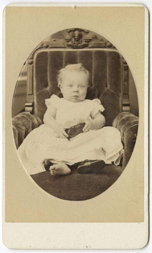 [Seated baby in armchair, holding one shoe and wearing another]