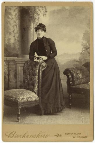[Woman standing against chair back, holding photograph]
