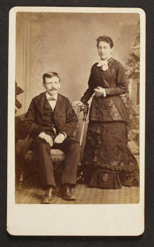 Unknown sitters [portrait of a standing woman and a seated man on upholstered chair]