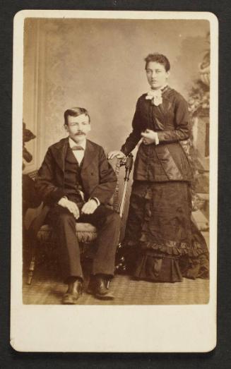 Unknown sitters [portrait of a standing woman and a seated man on upholstered chair]