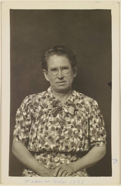 [Woman, seated, wearing patterned dress and wire glasses]