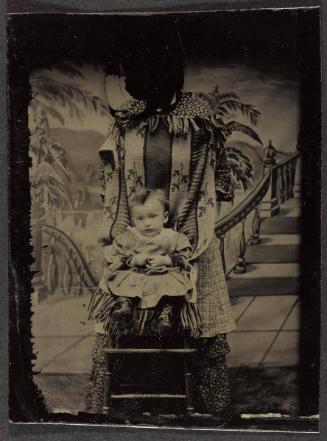 Portrait of young child seated in studio with painted backdrop (supported by shrouded figure)