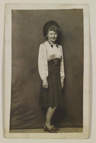 [Woman wearing dark skirt, silk shirt, hat, standing]