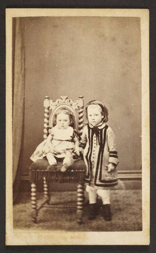 Unknown sitter [portrait of seated infant on upholstered chair left to a standing girl]