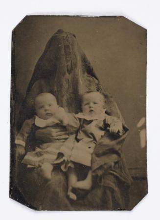 Portrait of twin babies (seated on lap of shrouded figure)
