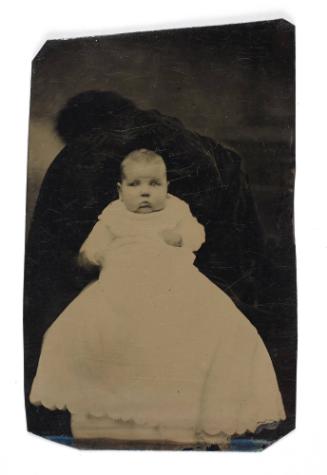Portrait of baby girl in white dress (seated on lap supported by defaced figure)